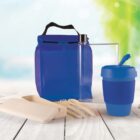 Reusable eco-friendly set with blue lunch bag, blue travel cup with straw, transparent water bottle, and white cutlery set with a beige container. Displayed on a wooden surface with a blurred green background.