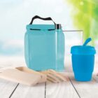Reusable lunch kit on a wooden surface featuring a blue insulated bag, transparent water bottle, blue cup with straw, and beige cutlery set with a matching container. The background is softly blurred greenery.