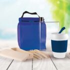 A reusable lunch set on a wooden surface includes a blue insulated bag, a transparent water bottle, a blue and white travel mug, a beige cutlery set with a case, and a bright outdoor background.
