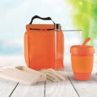 An orange insulated lunch bag with a clear water bottle, an orange travel mug, a beige cutlery set, and a beige food container. The background is blurred green and blue, suggesting an outdoor setting.