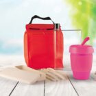 A reusable lunch set featuring a red insulated bag, a transparent water bottle, a pink travel mug with a straw, a set of beige utensils, and a matching container. The background is a blurred outdoor scene.