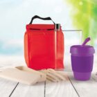 A red lunch bag, a clear water bottle, a purple reusable cup with a lid, a set of beige cutlery, and a beige case are displayed on a wooden surface. The background is a blurred outdoor scene with greenery and sunlight.