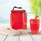 A red lunch bag with a black handle, a transparent water bottle, a red reusable cup with lid, eco-friendly cutlery set, and a biodegradable container are arranged on a white wooden table. The background is a blurred outdoor scene.