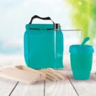 Reusable eco-friendly items on a wooden surface include a turquoise lunch bag, a transparent water bottle, a turquoise travel mug with a straw, a beige rectangular container, and beige cutlery. A blurred green background suggests an outdoor setting.