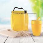 A reusable yellow lunch bag with a black handle is placed on a wooden surface. Next to it are a yellow travel cup, clear water bottle, beige cutlery set, and a beige container. The background shows a blurred green and blue outdoor scene.