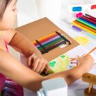 A child is drawing with colored pencils on a piece of paper at a table. The paper displays a colorful drawing. Various art supplies, including a set of colored pencils and a pair of 3D glasses, are scattered on the table.
