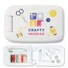 A sewing kit labeled Crafty Needles featuring scissors, buttons, and a spool of thread on the lid. Inside, the kit contains various thread colors, safety pins, buttons, a needle threader, and metal snap buttons, all neatly organized.