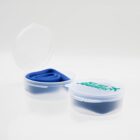 A small, transparent plastic container with a lid is open, revealing a folded blue silicone item inside. Another closed container is beside it, featuring a logo with the text Happy Journeys and an airplane graphic.