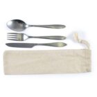 A spoon, fork, and knife made of metal are neatly arranged next to a beige fabric pouch with a drawstring. The pouch is positioned below the utensils.