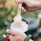 Two hands holding a bowl of rice, with one hand using a wooden spoon to scoop some rice. The spoon has fresh food printed on it in green. The background is blurred, showing outdoor greenery.