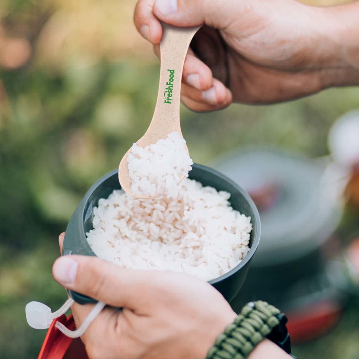 Two hands holding a bowl of rice, with one hand using a wooden spoon to scoop some rice. The spoon has fresh food printed on it in green. The background is blurred, showing outdoor greenery.