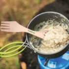 A camping pot on a portable stove contains oatmeal or porridge, with a wooden spork resting on the rim. The pot has green handles, and the background is a blurred natural outdoor setting.