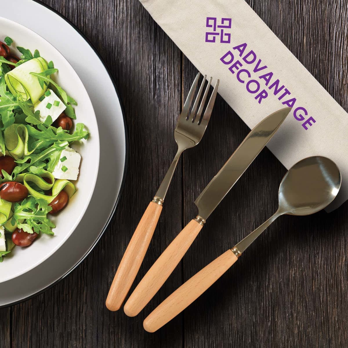 A salad with greens, zucchini ribbons, beans, and feta on a white plate beside a fork, knife, and spoon with wooden handles. A branded napkin with Advantage Decor text is under the utensils.