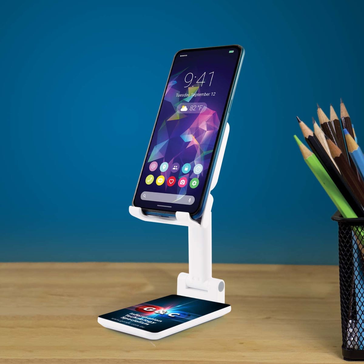 Smartphone on a white stand displaying a vibrant geometric wallpaper. A wireless charging pad below shows the word IGNC. A pencil holder with pencils is on the right. The background is a solid teal blue. The table is wooden.