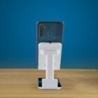 Smartphone placed vertically on a white stand, set on a wooden surface with a blue background. The phones rear features multiple camera lenses and a fingerprint sensor. The stand has an adjustable arm.