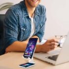 A person in a denim shirt is sitting at a desk, smiling. They are using a smartphone on a white stand in front of a laptop. The phone screen displays colorful app icons. An open notebook is visible next to the laptop.