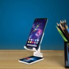 A smartphone rests on a white stand on a wooden surface, displaying a colorful screen with icons and the time 9:41. Next to it is a black mesh holder containing pencils. The background is a blue wall.