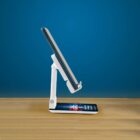 A smartphone on a white adjustable stand, angled upright against a blue background, placed on a wooden surface. The phone screen displays colorful graphics.
