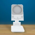A white, rectangular phone or tablet holder with an adjustable stand is placed on a wooden surface. The holder has a circular clamp and is positioned against a blue background.