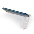 A blue smartphone is propped up at an angle on a white stand. The stand has two circular cutouts and is designed to hold the phone upright on a flat surface.
