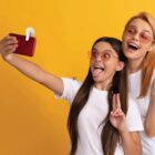 Two people wearing white shirts and orange-tinted sunglasses take a selfie against a yellow background. One person is holding the phone and making a peace sign while sticking out their tongue. Both are smiling.