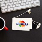 A white charging device with the Litewear Clothing logo, featuring orange, green, and yellow swooshes. It has USB and other connector ports. Its placed on a black surface with part of a keyboard and a red-handled coffee mug nearby.