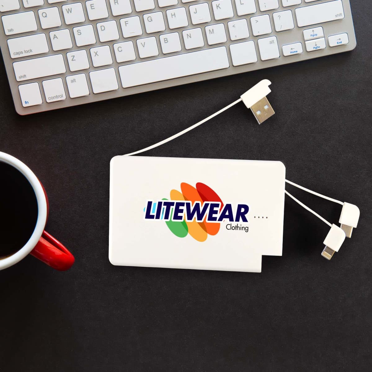 A white charging device with the Litewear Clothing logo, featuring orange, green, and yellow swooshes. It has USB and other connector ports. Its placed on a black surface with part of a keyboard and a red-handled coffee mug nearby.