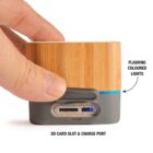 A hand holding a small bamboo Bluetooth speaker. It has a grey base featuring an SD card slot and charge port. There are flashing colored lights on the speakers side.