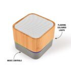 A small, square-shaped speaker with a bamboo top and perforated grille. The bottom is grey with music control buttons. Text annotates areas: Flashing coloured lights and Music controls.