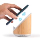 A hand is holding a smartphone above a wooden triangular-shaped speaker. Blue wireless signal waves are visible between the phone and the speaker, indicating a connection or interaction.