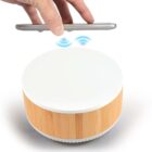 A hand holds a smartphone above a circular wooden device with a white top, illustrating wireless connection by emitting blue wave symbols.
