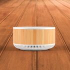 A round, light wood and white air purifier sits centrally on a wooden floor. The sleek design has a bamboo texture on the sides and a vented base, exuding a minimalist and modern aesthetic. The background features wooden planks.