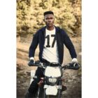 Man in a 17 shirt sitting on a dirt bike outdoors in a wooded area.