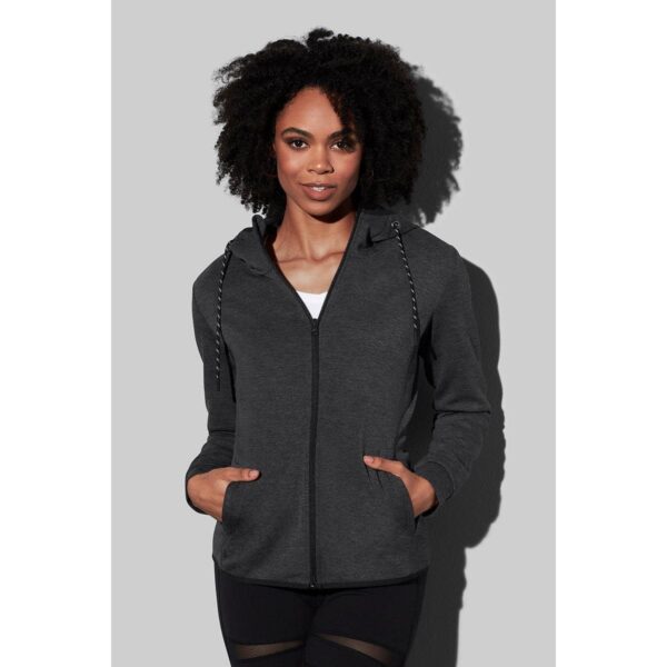Person wearing a dark gray zip-up hoodie with hands in pockets, standing against a gray background.