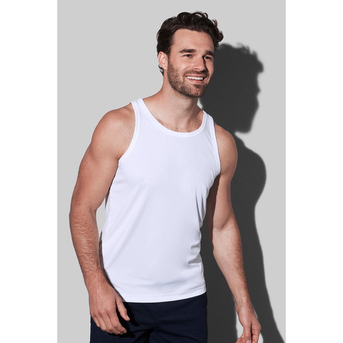 Man in a white tank top and dark pants stands against a gray background, smiling.