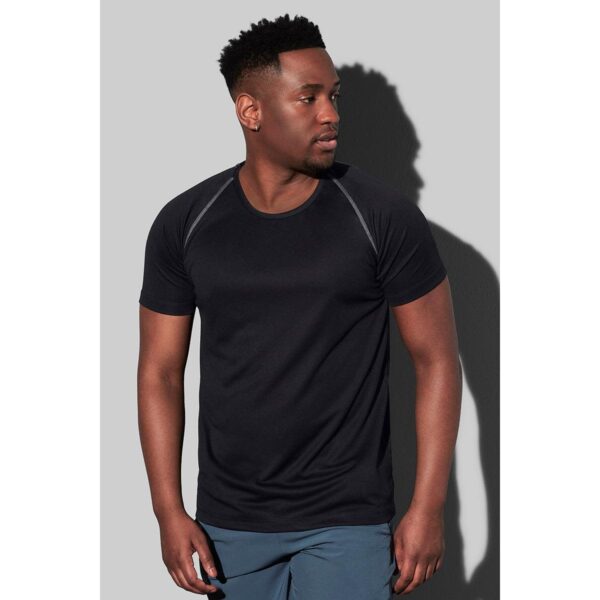Man in black T-shirt and blue shorts standing against a gray background.