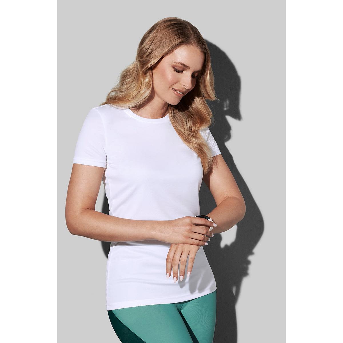 Woman in white t-shirt and green pants stands against a gray background, looking down and smiling.