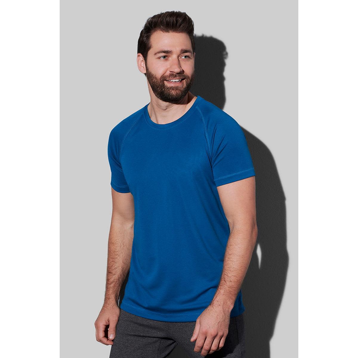 Man in blue T-shirt and jeans stands against a plain background.