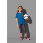 Girl in blue shirt holding a soccer ball, wearing leggings and sneakers, standing on gray background.