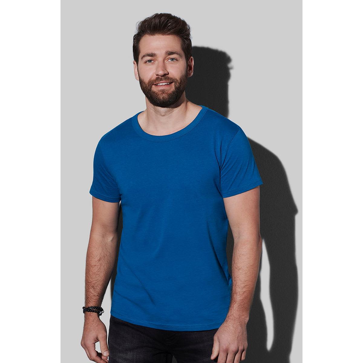 Man in a blue T-shirt and dark pants standing against a gray background.
