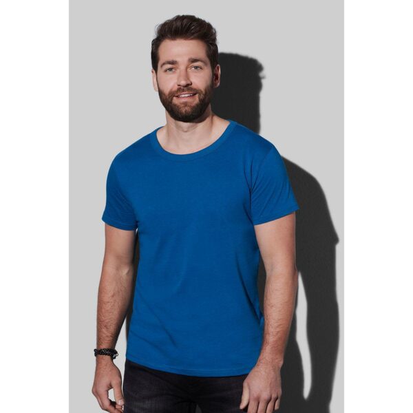 Man with a beard wearing a blue T-shirt and black jeans against a light gray background.