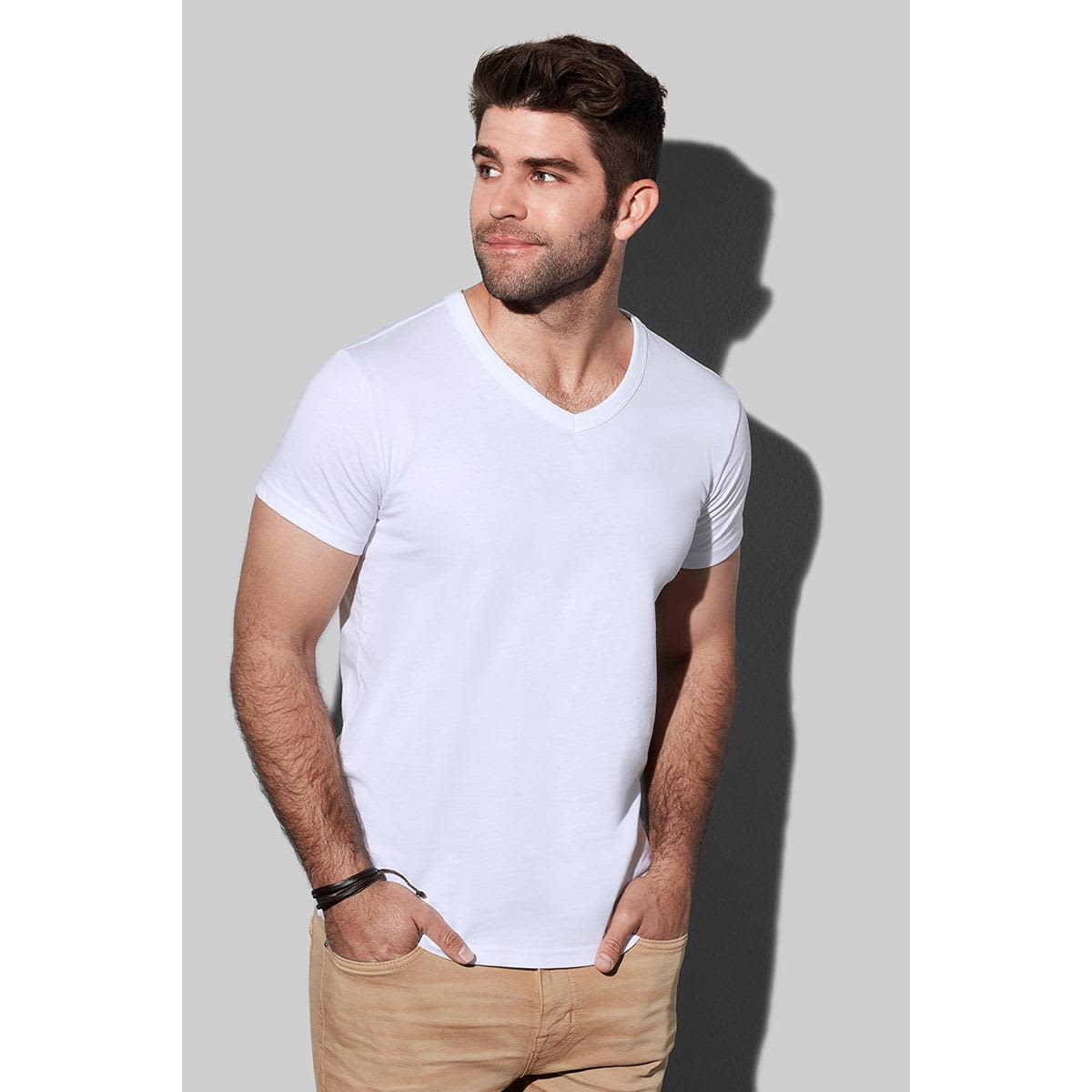 Man in a white V-neck T-shirt and khaki pants against a gray background.