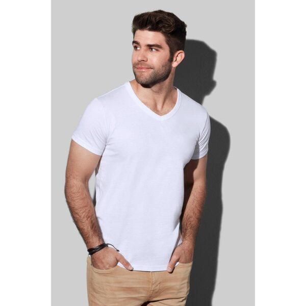 Man in a white V-neck shirt and beige pants standing against a gray background.