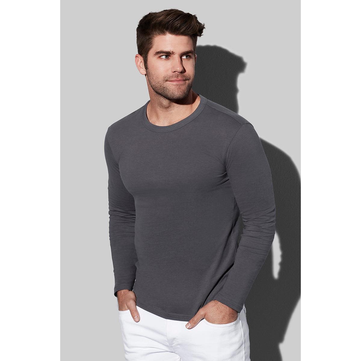 Man in gray long-sleeve shirt and white pants standing against a gray background.