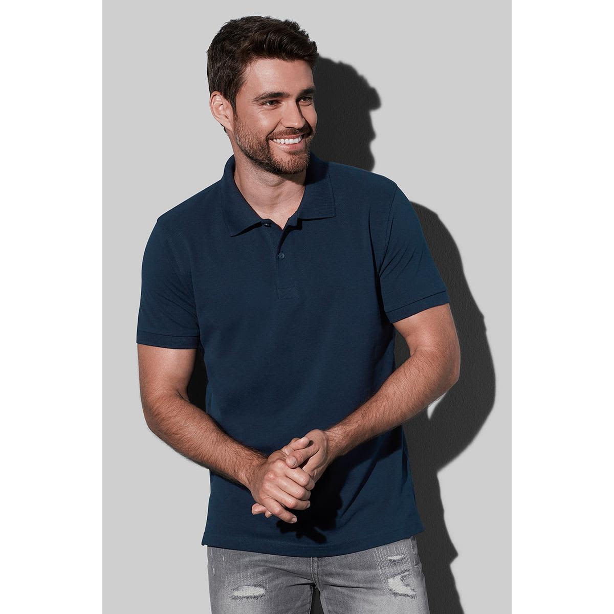 Smiling man in a dark polo shirt and jeans against a plain background.