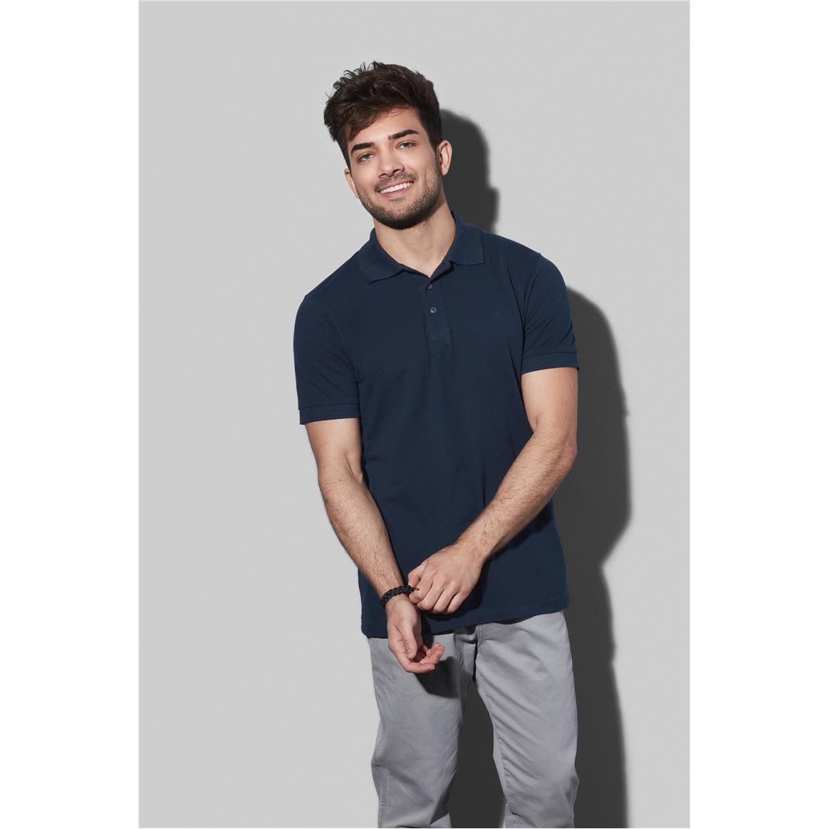 Smiling man in a navy polo shirt and gray pants stands against a light gray background.