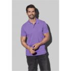 Smiling man wearing a purple polo shirt and black jeans against a gray background.