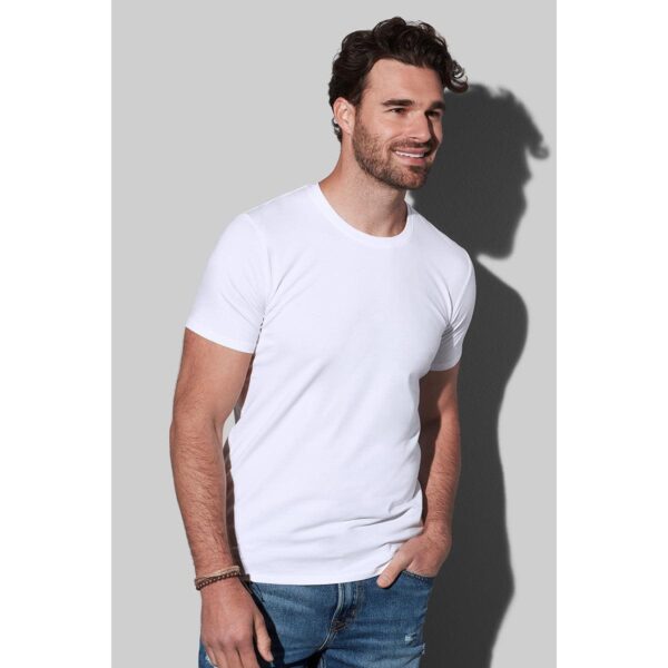 Man in white T-shirt and jeans smiling against a gray background.