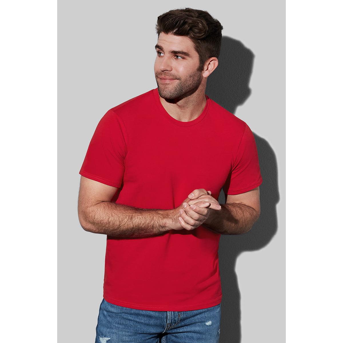 Man in a red T-shirt and jeans, standing against a gray background, with hands clasped.