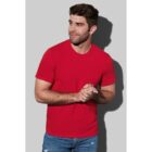 Man in red shirt and jeans stands against a gray background, looking to the side.